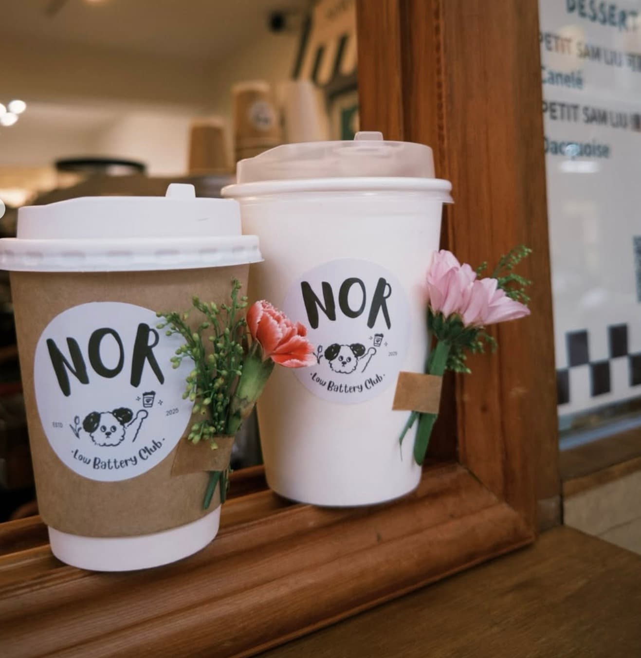 Two NOR Coffee cups with the Low Battery Club dog logo and fresh flowers, on the window counter